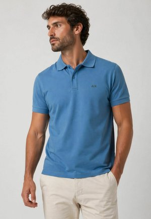 Uomo con polo blu a maniche corte e pantaloni beige, guarda a sinistra con una mano in tasca su sfondo semplice.