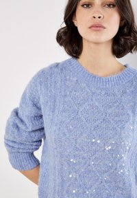 Hellblauer Pullover aus weichem, strukturiertem Material mit einem Zopfmuster und verstreuten Pailletten auf der Vorderseite. Verfügt über einen runden Halsausschnitt.
