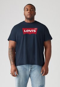 Tengerészkék pamut póló, középen nagy piros Levi's logóval, kerek nyakkal és rövid ujjal, világoskék, koptatott farmernadrággal párosítva.
