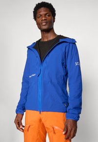 Sinine veekindel jakk kapuutsiga, ees lukk, elastsed mansetid ja logodetailidega. Kättevõetud erksate oranžide pükstega.