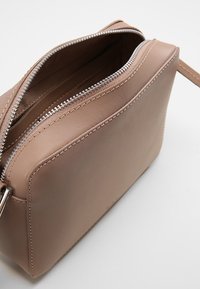 Borsa a tracolla in pelle beige con una texture liscia, chiusura con zip e dettagli di cucitura bianca. Forma rettangolare compatta con manico attaccato.
