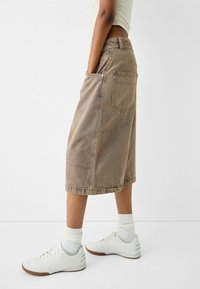 Jupe en denim marron atteignant le mollet, avec des poches latérales et une poche arrière, et une coupe décontractée. Associée à des baskets blanches et des chaussettes.