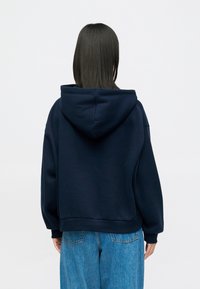 Άτομο με ίσιο σκούρο μαλλί, ντυμένο με ναυτίλο μπλε φού�τερ και μπλε τζιν, που στέκεται με την πλάτη στραμμένη προς μια λευκή απλή επιφάνεια.