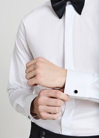 Camicia da uomo bianca con papillon nero, maniche lunghe e polsini con bottoni. Presenta gemelli neri a contrasto e una texture del tessuto liscia.