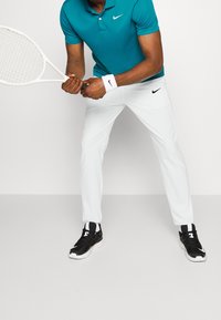 Türkises Nike-Polohemd, weiße Nike-Tennis-Hosen, schwarze Sportschuhe mit weißen Akzenten, hält einen weißen Tennisschläger.