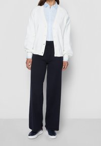Cardigan bianco con bottoni, indossato sopra una camicia azzurra chiara, abbinato a pantaloni a gamba larga blu navy e sneakers blu navy.