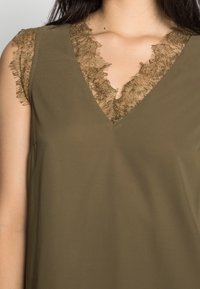 Olivi vihreä paita, jossa on V-pääntie, kauniisti frayed-kultalankareunus sekä lyhyet, pitsiaksentilla varustetut hihat.