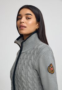 Grå cable-knit tröja med hög krage, utrustad med en svart dragkedja och en logopatch på vänster ärm. Slät textur och figurnära design.