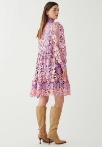 Nėriniuota suknelė purpuro ir rožinės spalvos gėlių raštu, turinti ilgus rankovėmis ir išplatintą sijoną. Dėvima su kelio aukščio tan spalvos batais.