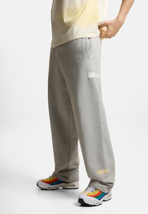 Osoba nosząca luźne szare spodnie dresowe z przetarciami oraz białe buty sportowe z żółtymi, czerwonymi, niebieskimi i zielonymi paskami, stojąca na białym tle.