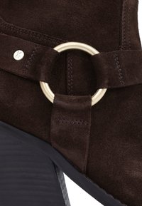 Brown suede ankle boot featuring a gold metal ring detail and a textured black heel. Stitching visible around the strap.
