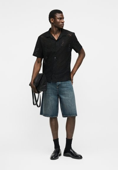 Άνδρας που φοράει μαύρο δαντελένιο πουκάμισο με κοντά μανίκια, τζιν σορτς μέχρι το γόνατο, μαύρες κάλτσες και παπούτσια, κρατώντας μαύρη τσάντα, κοιτάζει προς τα πλάγια.