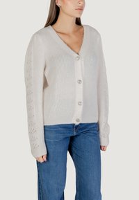 Cardigan de punto en color crema con escote en V, cinco botones y patrones de manga calados. Textura suave y corte holgado. Combinado con jeans azules.