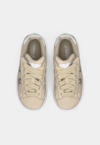 Bézs színű velúr sportcipők fehér gumitalppal, lapos bézs cipőfűzőkkel, oldalsó csíkokkal és egy fémes díszítéssel a nyelvén.