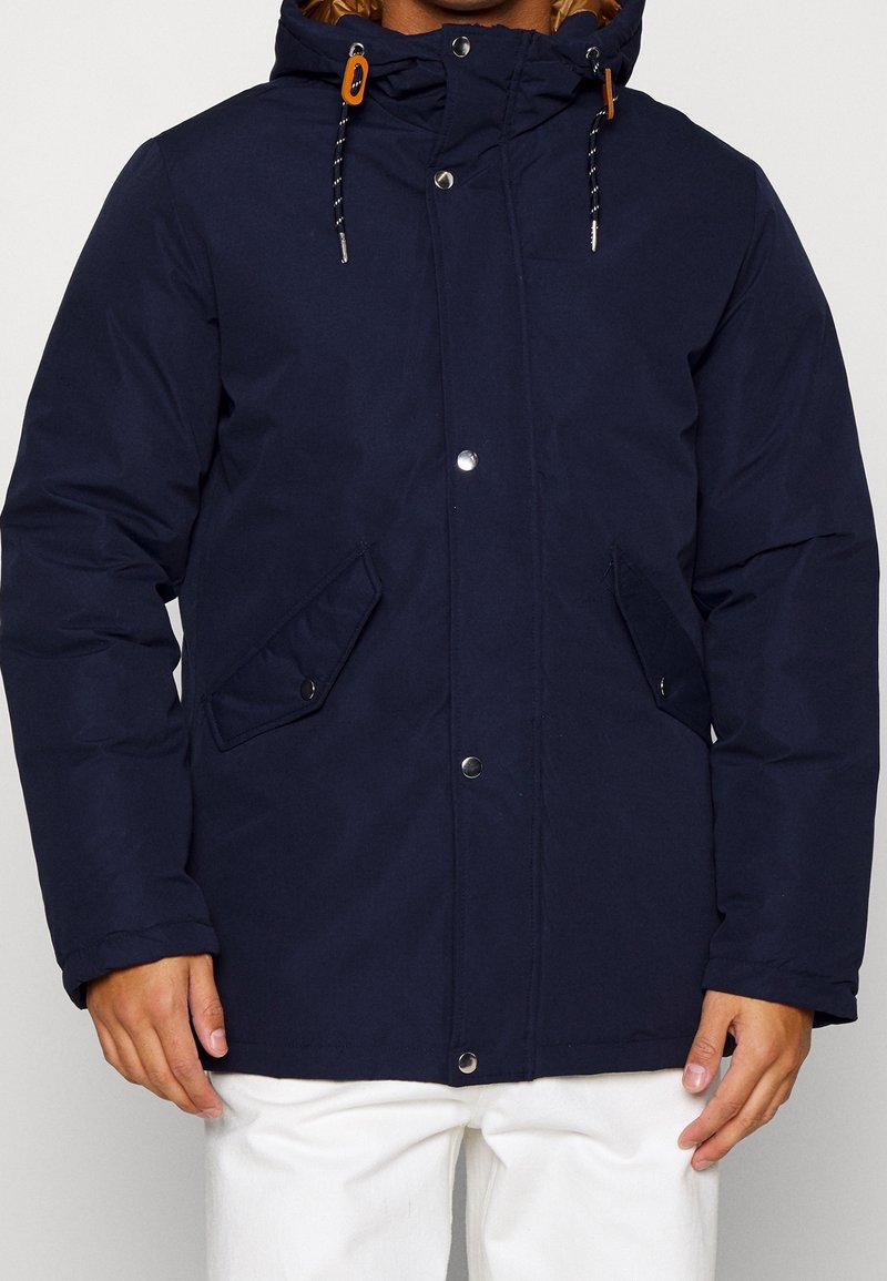 Marinblå isolerad jacka med dragkedja och tryckknappar, justerbar huva, två fickor på sidan och kontrasterande orangea detaljer på dragsnören.