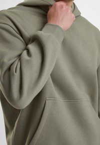 Felpa con cappuccio verde oliva realizzata in cotone morbido, con tasca a marsupio e polsini a costine. Il tessuto ha una texture liscia con cuciture minimali.