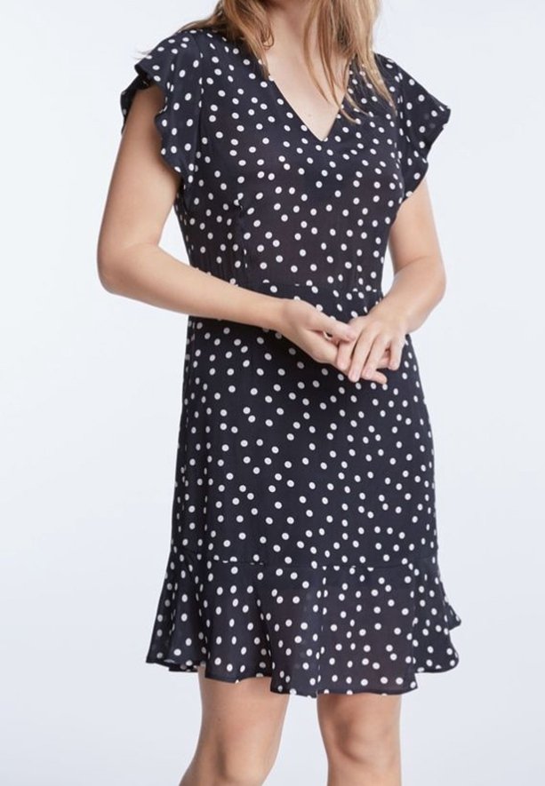 Femme portant une robe noire à manches courtes avec des pois blancs et un ourlet à volants, debout, les mains jointes devant elle.