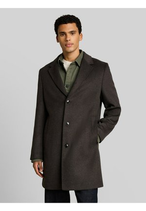 Homme portant un manteau marron foncé boutonné sur une chemise verte, debout avec une main dans la poche devant un fond gris uni.
