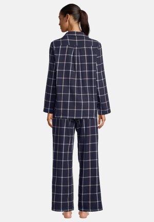 Donna in piedi a piedi nudi che indossa un pigiama blu navy con motivo a quadri bianchi, vista da dietro su sfondo bianco.