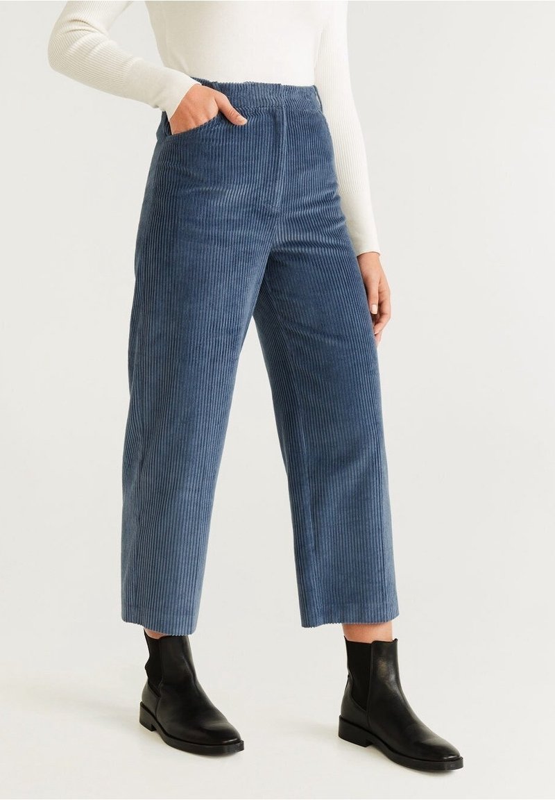 Femme portant un pantalon large en velours côtelé bleu, un haut blanc à manches longues et des bottines noires, debout avec une main dans la poche.