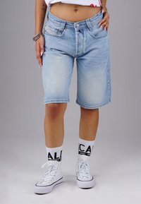 Helle blaue Denim-Shorts mit lockerer Passform, ausgefranster Saum und zwei vorderen Taschen. Kombiniert mit weißen High-Top-Sneakern und gemusterten Socken.