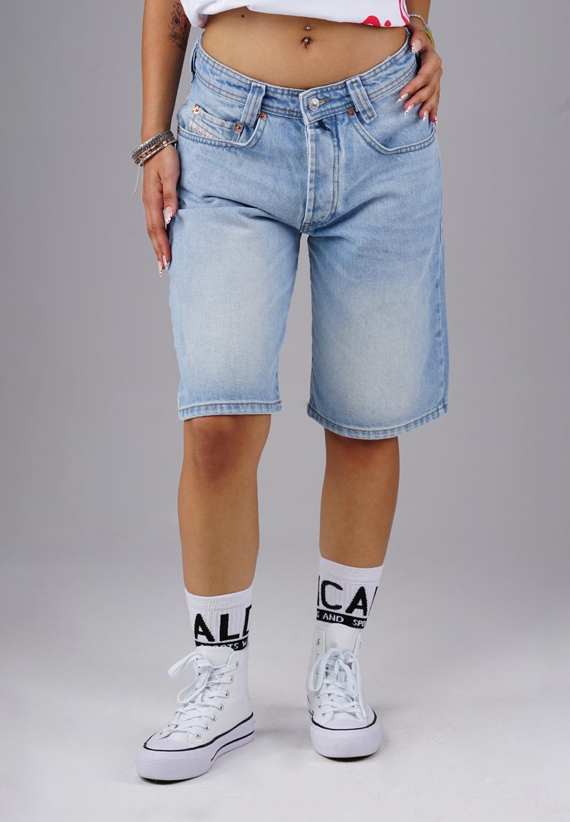 Helle blaue Denim-Shorts mit lockerer Passform, ausgefranster Saum und zwei vorderen Taschen. Kombiniert mit weißen High-Top-Sneakern und gemusterten Socken.