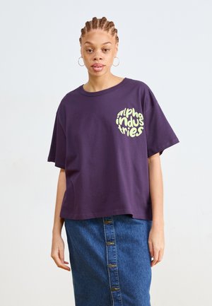 Fialové bavlnené tričko s voľným strihom, okrúhlym výstrihom a krátkymi rukávmi. Na prednej strane má žlté kruhové logo.