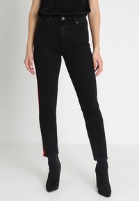 Černé džínsy s vysokým pasem, úzkého střihu, s červeným bočním pruhem a klasickým pětiokou designem. Materiál vypadá elasticky, s jemnou texturou.