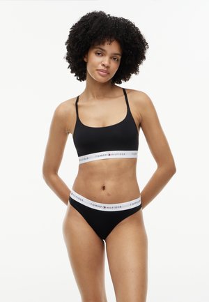 Γυναίκα με σγουρά μαλλιά που φοράει μαύρο bralette Tommy Hilfiger και ταιριαστά εσώρουχα με λευκές ελαστικές ταινίες με λογότυπο, στέκεται μπροστά σε λευκό φόντο.