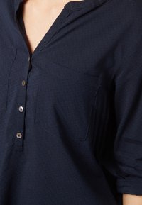 Chemise bleu marine à boutons en tissu léger, avec une poche sur la poitrine gauche et un motif texturé. Les boutons sont de couleur argentée.