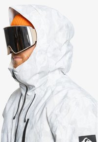 Weiße Jacke im Camouflage-Muster mit hohem Kragen, verstellbarem Kapuze und schwarzem Reißverschluss. Ausgestattet mit großen, spiegelnden Goggles und einer orangefarbenen Strickmütze.