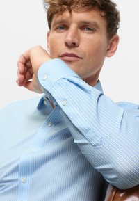 Man wearing light blue and white striped button-up shirt resting arm on chair, looking forward with neutral expression.