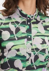 Donna che indossa una camicetta button-up a righe verdi e nere con colletto arricciato e motivi di foglie astratte.