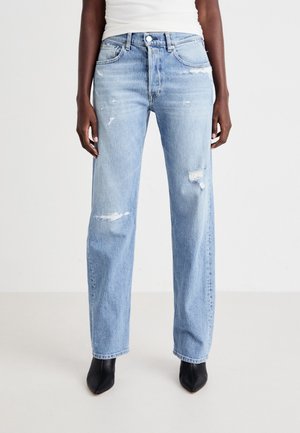 Personne portant un jean droit délavé bleu clair, un haut blanc et des bottines noires à bout pointu, debout devant un fond uni.