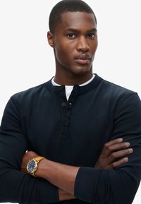 Young man with crossed arms, wearing a navy buttoned long-sleeve shirt and a gold watch with a brown leather strap.