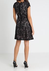 Schwarzes Spitzenkleid mit kurzen Cap-Ärmeln, floralem Muster, ausgestelltem Saum und offenem Reißverschluss am Rücken. Kombiniert mit schwarzen High Heels.