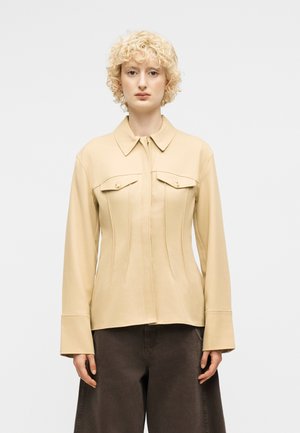 Ξανθός με κυματιστά μαλλιά που φοράει μπεζ μακρυμάνικο πανωφόρι με γιακά και φερμουάρ, με τσέπες στο στήθος με κουμπιά, και σκούρο παντελόνι με φαρδιά μπατζάκια.