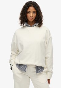 Sudadera crema de corte relajado, con escote en V y puños de canalé, superpuesta a una camisa de rayas en blanco y negro. Textura suave.