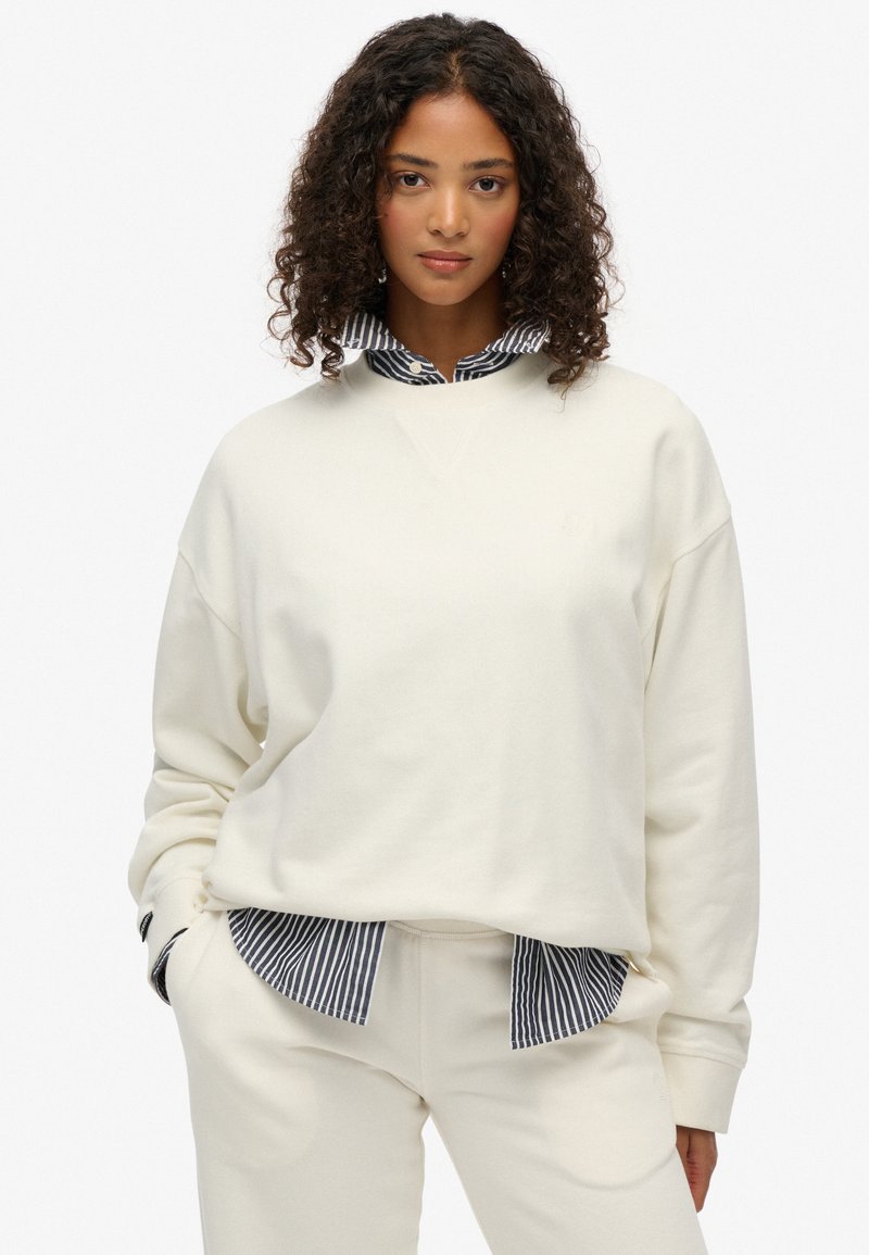 Sudadera crema de corte relajado, con escote en V y puños de canalé, superpuesta a una camisa de rayas en blanco y negro. Textura suave.