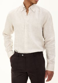 Camicia in lino beige chiaro con colletto button-down, maniche lunghe e una texture sottile, abbinata a pantaloni marroni scuri con chiusura a bottone.
