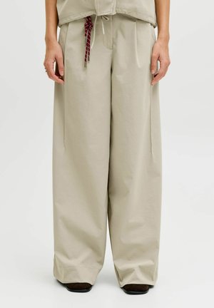 Bézs, bőszárú nadrág piros-fehér zsinórhúzóval, hozzáillő felsővel és fekete cipővel, sima háttér előtt állva.