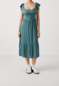 Tyrkis knälång klänning med puffärmar, rynkat midja och utsvängd kjol. Matchas med vita sneakers och grå strumpor.