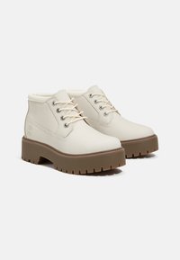 Beige nilkkurit, jotka on valmistettu sileästä nahasta, beigeillä nauhoilla, pyöreällä varvalla, pehmustetulla nilkkareunalla ja paksulla ruskealla kumipohjalla, jossa on urat.