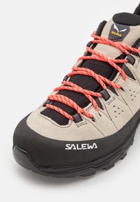 Chaussure de randonnée en daim beige avec des accents noirs, des lacets orange vif et une semelle en caoutchouc noire. Présente des motifs triangulaires uniques sur les côtés.