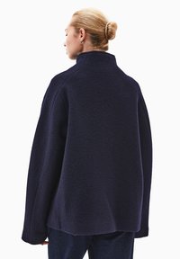 Przesadny granatowy płaszcz z teksturowanej wełny, z wysokim kołnierzem, opadającymi ramionami i szerokimi rękawami, widziany z tyłu.