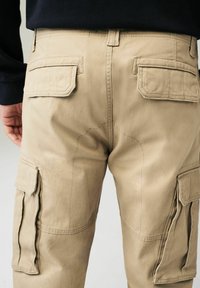 Pantaloni cargo bej cu două buzunare la spate, buzunare laterale cargo și detalii de cusătură vizibile. Fabricat dintr-un material durabil cu o textură netedă.