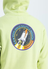 Svetlozelená mikina s grafikou zobrazujúcou raketoplán, hviezdne vzory a text "100. misia raketoplánu".