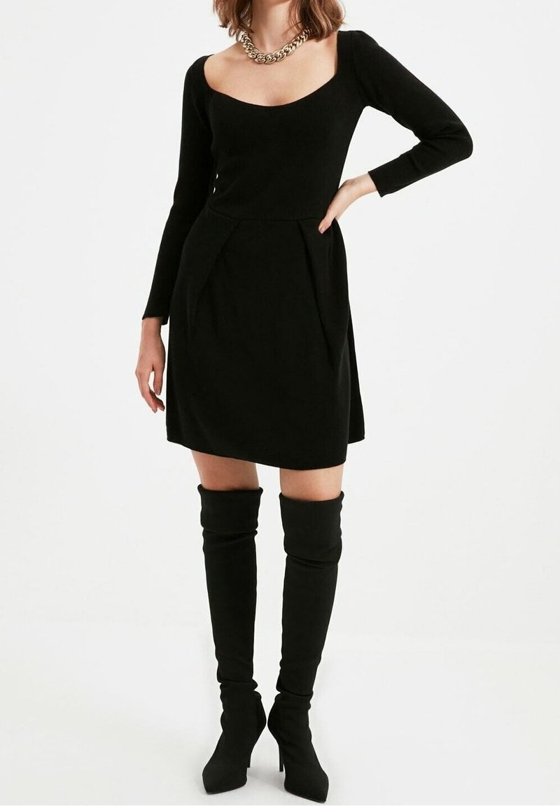 Black long-sleeve dress with a scoop neckline, pleats at the waist, and a fitted design. Paired with thigh-high black boots.