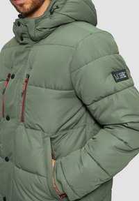 Groene pufferjack met een capuchon, voorzien van rode rits zakken, zwarte knopen en een patch op de mouw. Gladde textuur en gestepte design.