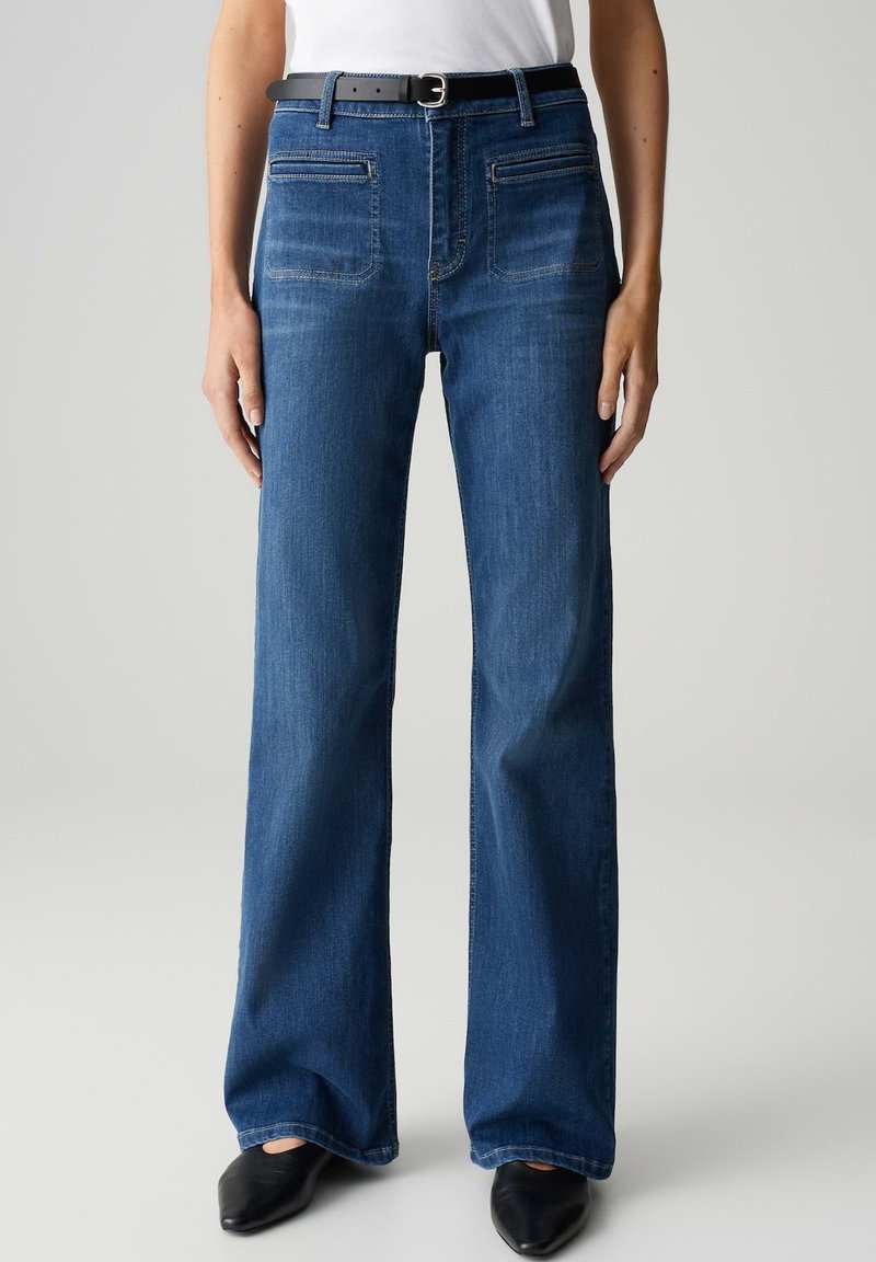 Brede spijkerbroek in blauw denim met een hoge taille, twee voorzakken en een zwarte riem, gedragen met zwarte puntige schoenen. Glad textuur.